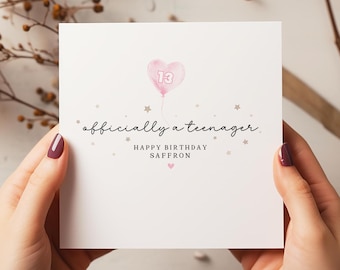 May include: A white card with a pink heart-shaped balloon with the number 13 inside. The text "officially a teenager" is written in black cursive. The text "Happy Birthday Saffron" is written in black below the balloon.