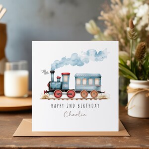 May include: A birthday card with a watercolour illustration of a blue train with red wheels and blue smoke. The card reads "Happy 2nd Birthday Charlie" in black and grey text. The card is on a brown envelope.