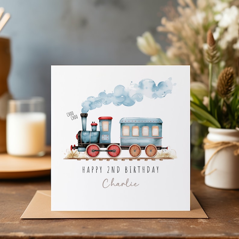 May include: A birthday card with a watercolour illustration of a blue train with red wheels and blue smoke. The card reads "Happy 2nd Birthday Charlie" in black and grey text. The card is on a brown envelope.