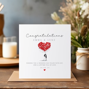 May include: A white card with the text "Congratulations Emma & Luke" and a drawing of a bride and groom holding red heart balloons. The text "Wishing you a wedding day filled with joy. And a lifetime full of love 30.08.24" is also on the card.