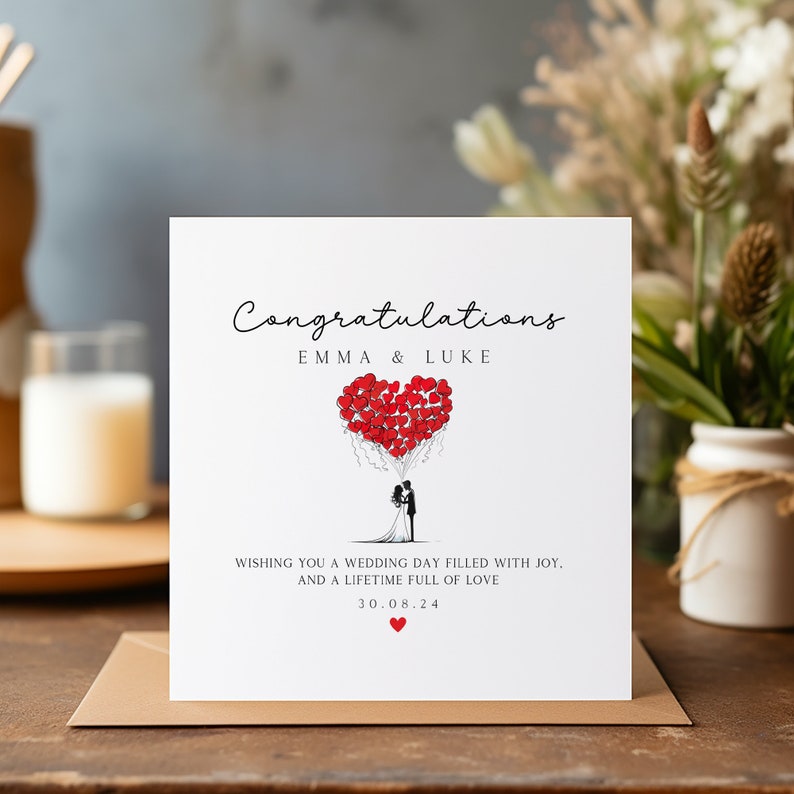 May include: A white card with the text "Congratulations Emma & Luke" and a drawing of a bride and groom holding red heart balloons. The text "Wishing you a wedding day filled with joy. And a lifetime full of love 30.08.24" is also on the card.