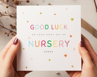 May include: A white greeting card with the words "GOOD LUCK" and "NURSERY" in colorful letters. The card also includes the text "ON YOUR FIRST DAY OF" and the name "POPPY". Small star illustrations are scattered around the text.