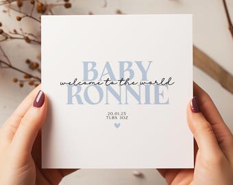 May include: A white card held by hands, featuring the text "BABY RONNIE welcome to the world" in blue and black script. The card also includes the date 20.01.25, weight 7lbs 3oz, and a small heart symbol.
