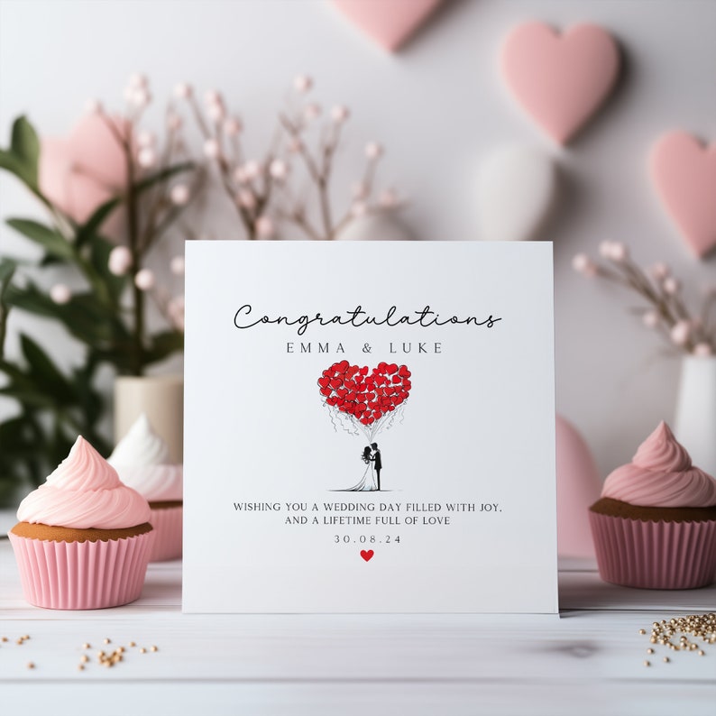May include: A white card with the text "Congratulations Emma & Luke" and a drawing of a bride and groom holding red heart-shaped balloons. The text below the drawing reads "Wishing you a wedding day filled with joy. And a lifetime full of love 30.08.24".