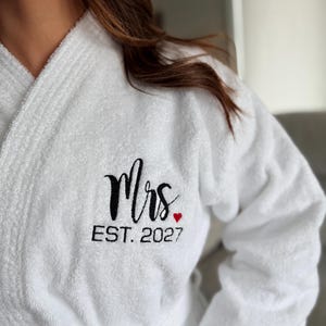 May include: White terry cloth robe with the embroidered word "Mrs" in black script, a small red heart, and "EST. 2027" in black block letters. The robe has a shawl collar and a belt.
