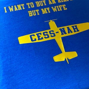 Peut inclure: Un t-shirt bleu vif avec un texte jaune qui dit I WANT TO BUY AN AIRPLANE BUT MY WIFE. Sous le texte se trouve un graphique jaune d'un avion avec le texte bleu CESS-NAH imprim&eacute; sur les ailes de l'illustration de l'appareil.