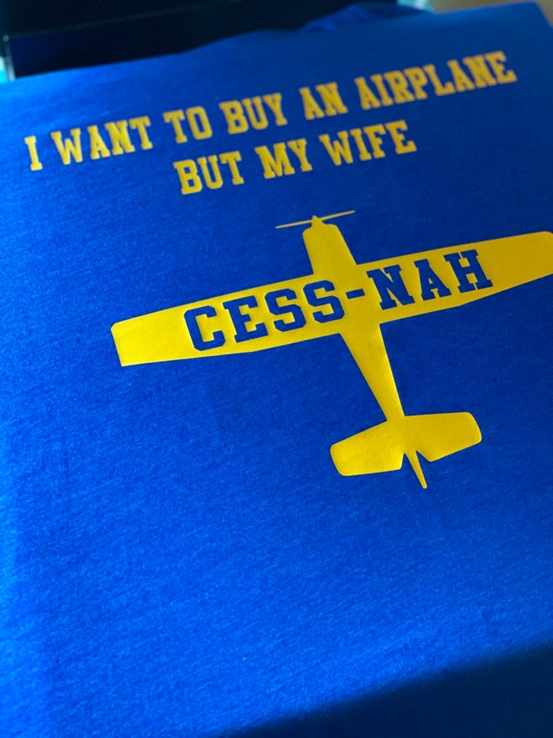 Peut inclure: Un t-shirt bleu vif avec un texte jaune qui dit I WANT TO BUY AN AIRPLANE BUT MY WIFE. Sous le texte se trouve un graphique jaune d'un avion avec le texte bleu CESS-NAH imprim&eacute; sur les ailes de l'illustration de l'appareil.