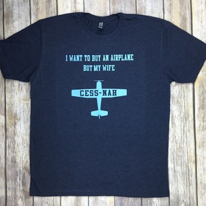 Peut inclure: Un t-shirt bleu marine &agrave; manches courtes avec un texte bleu clair indiquant I WANT TO BUY AN AIRPLANE BUT MY WIFE et un graphique d'avion avec le texte CESS-NAH imprim&eacute; sur les ailes dans une police de couleur bleu marine fonc&eacute;.
