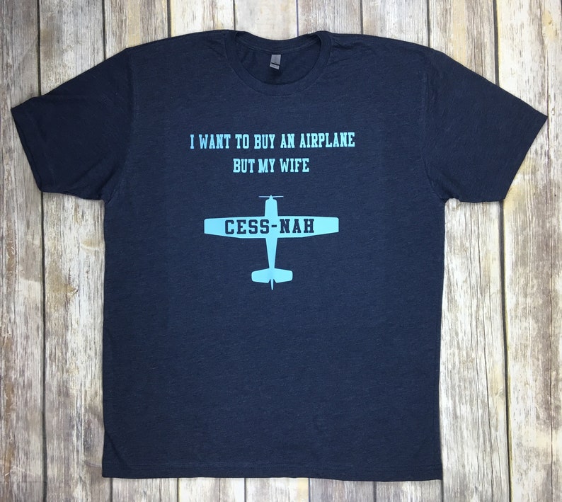 Peut inclure: Un t-shirt bleu marine &agrave; manches courtes avec un texte bleu clair indiquant I WANT TO BUY AN AIRPLANE BUT MY WIFE et un graphique d'avion avec le texte CESS-NAH imprim&eacute; sur les ailes dans une police de couleur bleu marine fonc&eacute;.