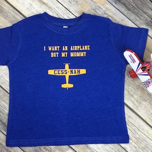 Peut inclure: Un t-shirt bleu roi &agrave; manches courtes avec le texte jaune I WANT AN AIRPLANE BUT MY MOMMY et un graphique d'avion jaune avec le texte CESS-NAH sur les ailes. Un petit avion jouet rouge, blanc et bleu est plac&eacute; sur le c&ocirc;t&eacute; droit.