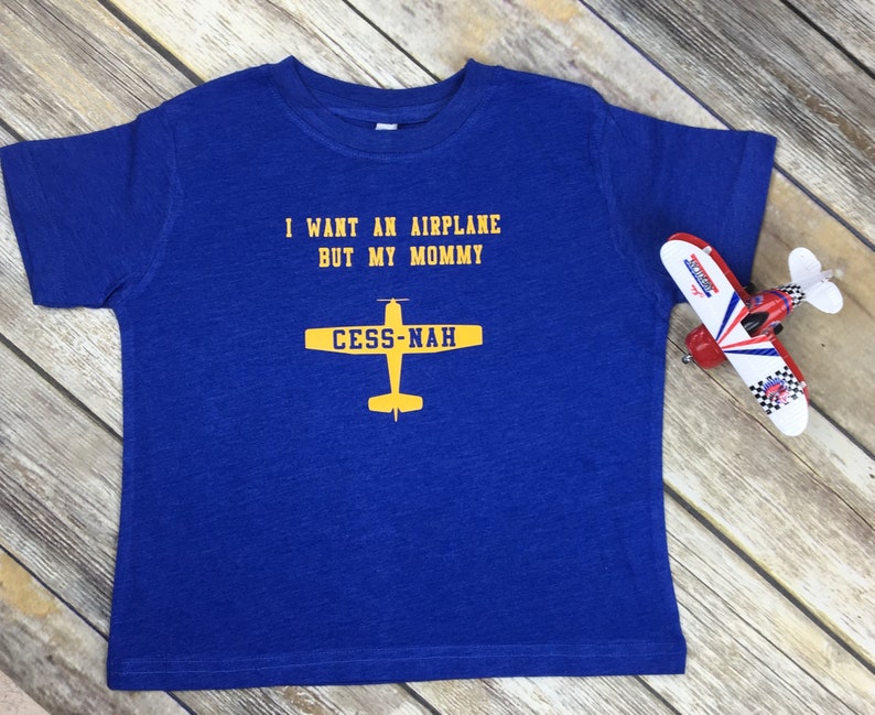 Peut inclure: Un t-shirt bleu roi &agrave; manches courtes avec le texte jaune I WANT AN AIRPLANE BUT MY MOMMY et un graphique d'avion jaune avec le texte CESS-NAH sur les ailes. Un petit avion jouet rouge, blanc et bleu est plac&eacute; sur le c&ocirc;t&eacute; droit.