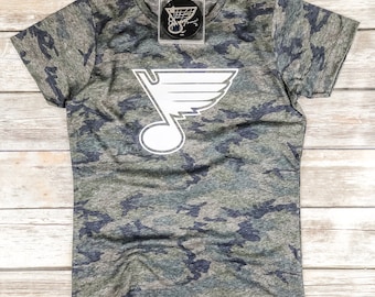 Può includere: Una t-shirt a maniche corte con motivo mimetico verde, grigio e nero e un grande logo bianco a forma di nota musicale dei St. Louis Blues centrato sul petto. Un disco da hockey nero con un logo bianco è posizionato vicino allo scollo.