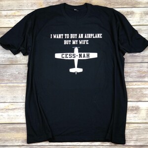 Peut inclure: Un t-shirt noir &agrave; manches courtes avec le texte blanc I WANT TO BUY AN AIRPLANE BUT MY WIFE et un graphique blanc repr&eacute;sentant la silhouette d'un avion avec le texte CESS-NAH imprim&eacute; sur les ailes du design de l'appareil.