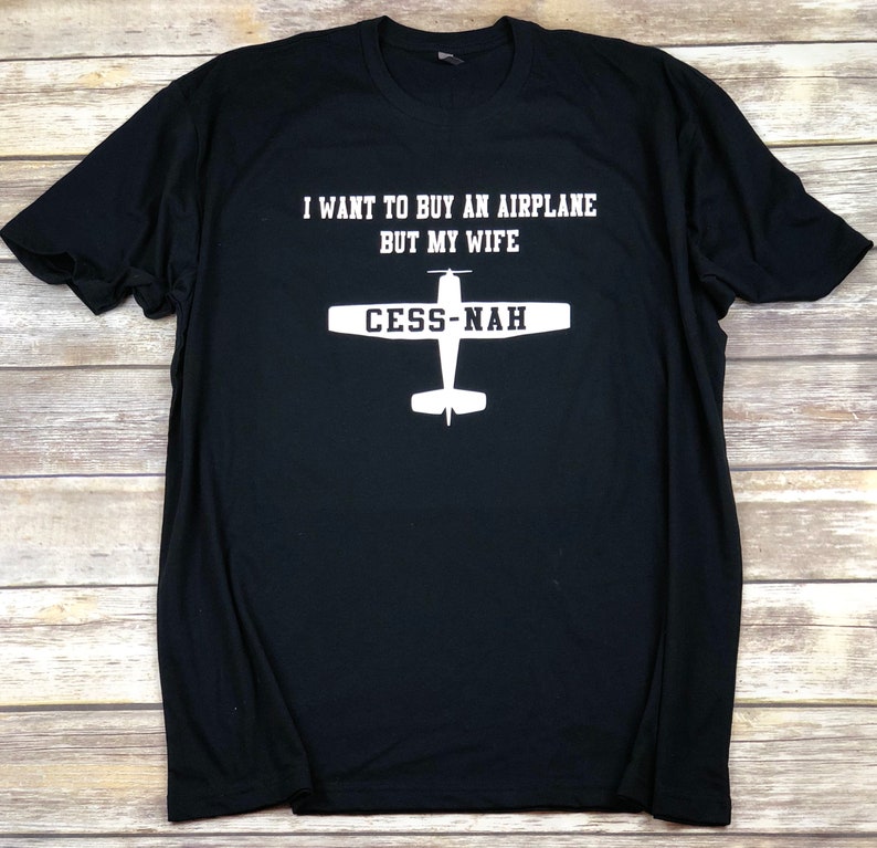 Peut inclure: Un t-shirt noir &agrave; manches courtes avec le texte blanc I WANT TO BUY AN AIRPLANE BUT MY WIFE et un graphique blanc repr&eacute;sentant la silhouette d'un avion avec le texte CESS-NAH imprim&eacute; sur les ailes du design de l'appareil.