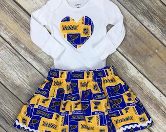 Può includere: Un body bianco a maniche lunghe con una toppa a forma di cuore che mostra il logo e la scritta dei St. Louis Blues, abbinato a una gonna coordinata con motivo a griglia blu e giallo, logo della squadra, scritta e orlo con bordo ondulato bianco.