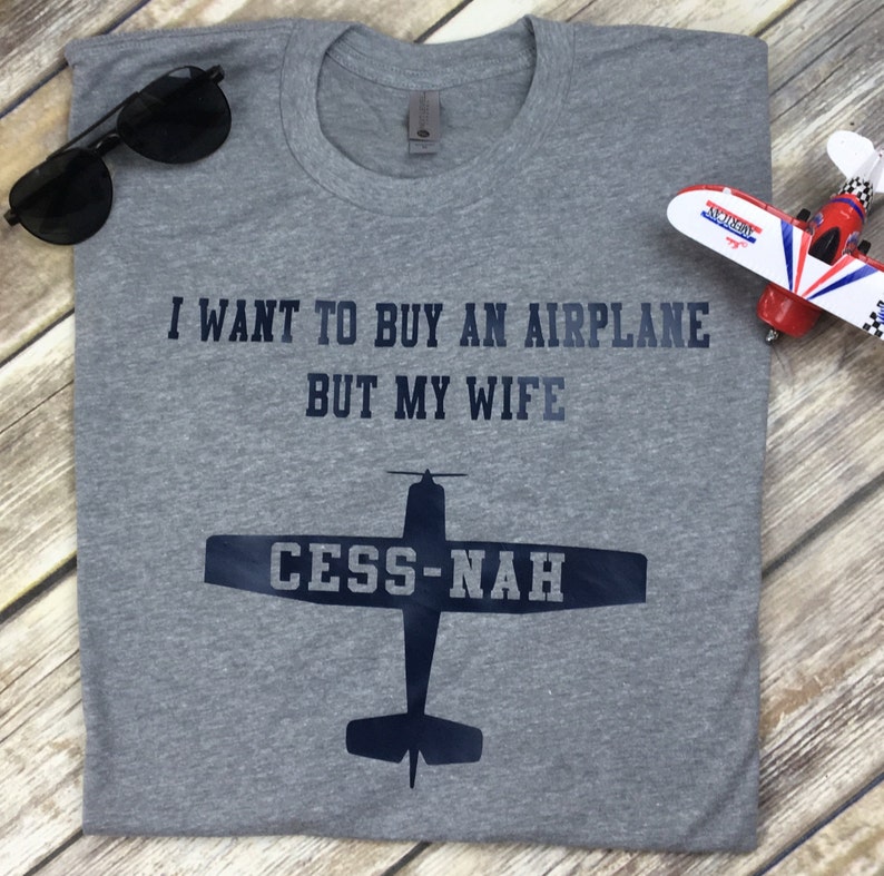 Peut inclure: Un t-shirt gris chin&eacute; avec le texte bleu fonc&eacute; I WANT TO BUY AN AIRPLANE BUT MY WIFE et un graphique repr&eacute;sentant la silhouette d'un avion avec le texte CESS-NAH sur les ailes. Le t-shirt est accompagn&eacute; de lunettes de soleil et d'un avion jouet.