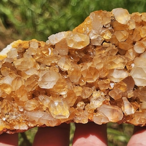 Puede incluir: Primer plano de un grupo de cristales de citrino dorado. Los cristales son una mezcla de tonos ámbar claros y oscuros, con una superficie rugosa y texturizada. Los cristales están agrupados, creando un efecto brillante. El fondo está borroso.