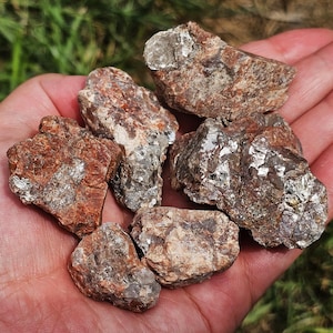 Könnte beinhalten: Eine Handvoll rauer, rotbrauner Steine mit glitzernden, silbernen Einschlüssen. Die Steine variieren in Größe und Form und haben ein natürliches, unpoliertes Aussehen. Der Hintergrund zeigt grünes Gras.