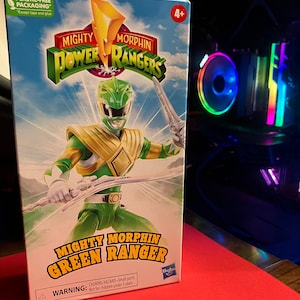 Può includere: Una action figure verde dei Power Rangers in una scatola. La figura tiene una spada e indossa un elmetto verde con una visiera dorata. La scatola è verde e bianca e ha le parole "Mighty Morphin Green Ranger" su di essa.