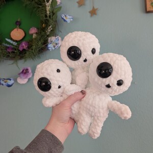 A bundle of three crocheted kodomas from the film “Princess Mononoke” against a blue wall background. They feature three different safety eye sizes. One large eye, one medium, and a small mouth.