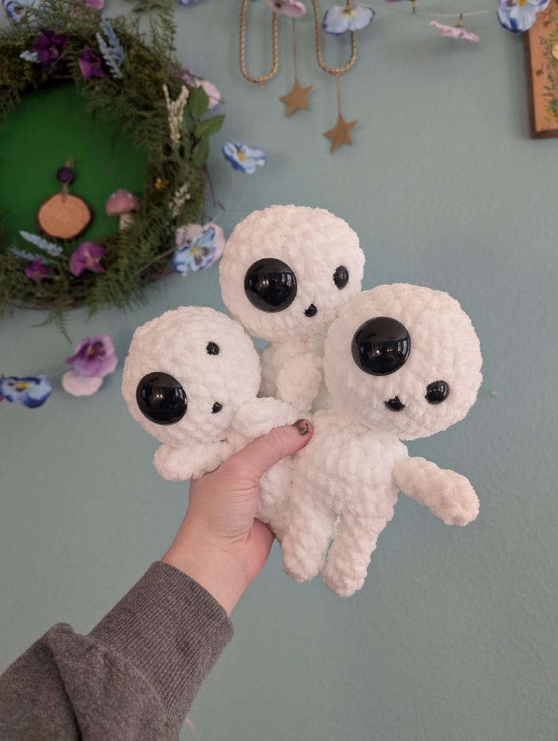 A bundle of three crocheted kodomas from the film “Princess Mononoke” against a blue wall background. They feature three different safety eye sizes. One large eye, one medium, and a small mouth.