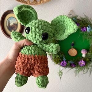 May include: A handmade green crochet doll with large black eyes and large ears. The doll is wearing brown shorts. The doll is a character from a popular science fiction franchise. The doll is approximately 8 inches tall.
