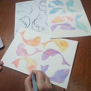 May include: A person paints watercolor fish on white paper. The fish are in shades of orange, pink, purple, and blue. The person is using a gray paintbrush.