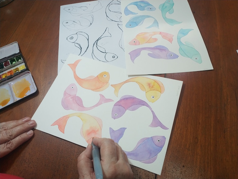 May include: A person paints watercolor fish on white paper. The fish are in shades of orange, pink, purple, and blue. The person is using a gray paintbrush.