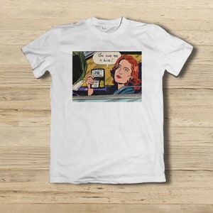 Può includere: Una t-shirt bianca con un'immagine in stile fumetto di una donna con i capelli rossi che tiene un distintivo dell'FBI e dice "You owe me a kiss!"