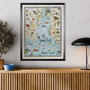 May include: Framed map print of Mobile Bay and the Gulf of Mexico, featuring detailed illustrations of local wildlife, landmarks, and nautical elements. The map is in a black frame and hangs above a wooden cabinet.
