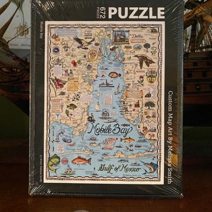 May include: A packaged puzzle featuring a detailed map of Mobile Bay, with illustrations of local landmarks, fish, and wildlife. The puzzle has 672 pieces and is labeled "Custom Map Art By Melissa Smith."