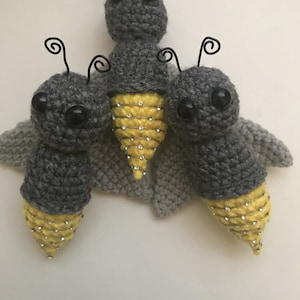 May include: Three gray crocheted fireflies with yellow and gold beaded bodies. The fireflies have black wire antennae and black eyes.