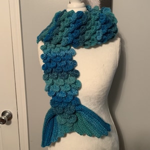 May include: A blue and green crocheted mermaid tail scarf. The scarf is made with a textured stitch that resembles scales.