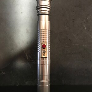 May include: A silver lightsaber with a gold top and a red button. The lightsaber has a textured surface and a black handle.