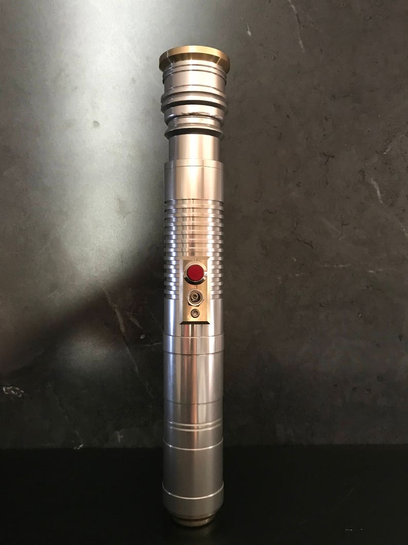 May include: A silver lightsaber with a gold top and a red button. The lightsaber has a textured surface and a black handle.