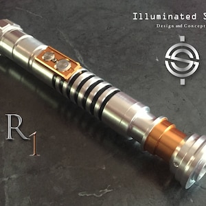 Può includere: Un'arma di scena di spada laser argentata e dorata con accenti neri e una punta color rame. La spada laser è etichettata "R1" e "Illuminated Saber Design and Concepts".