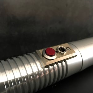 May include: A silver lightsaber with a red button and a black power cord. The lightsaber has a textured grip.
