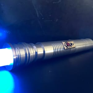 May include: A silver lightsaber with a blue blade. The lightsaber has a red button and a silver handle with a textured design.