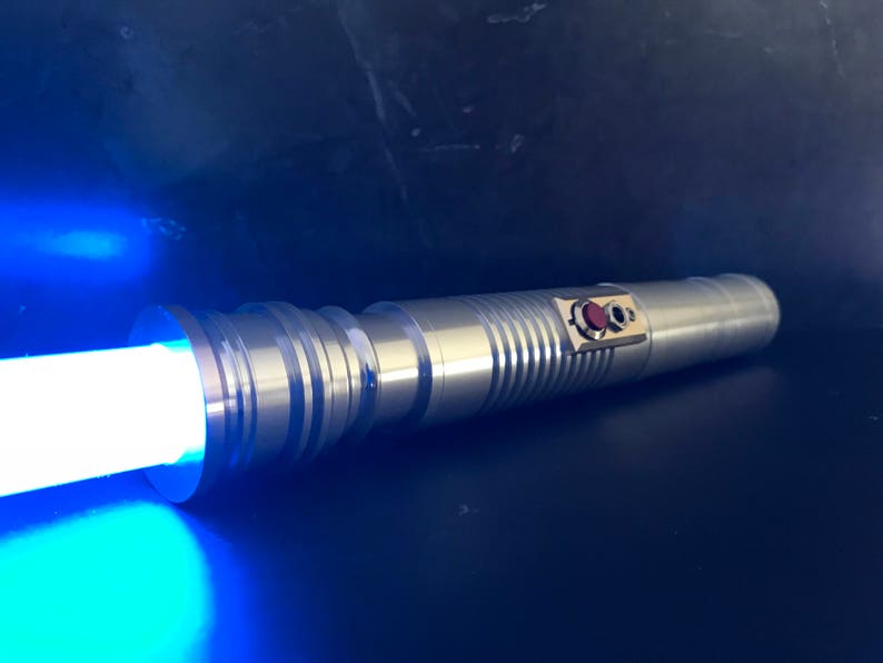 May include: A silver lightsaber with a blue blade. The lightsaber has a red button and a silver handle with a textured design.