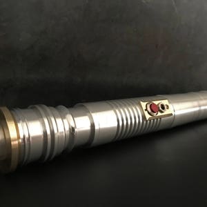 May include: A silver lightsaber with a brass ring at the end and two red buttons on the side.