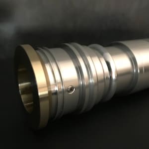 May include: A silver lightsaber hilt with a gold ring around the end. The hilt has a textured surface and a small screw visible on the side.