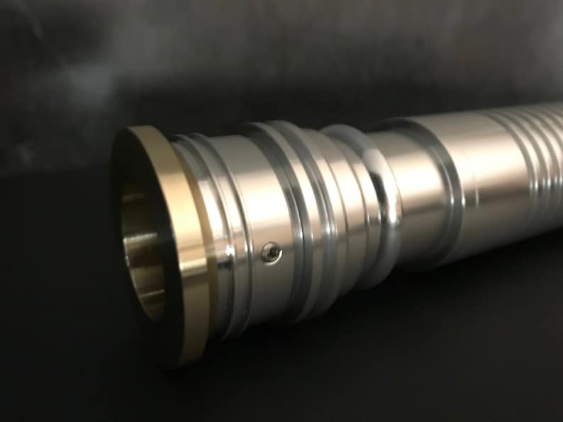 May include: A silver lightsaber hilt with a gold ring around the end. The hilt has a textured surface and a small screw visible on the side.