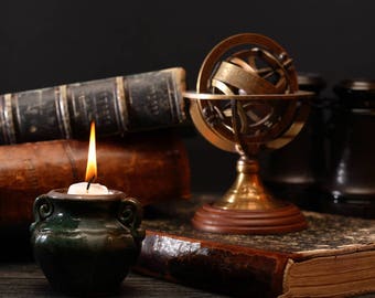 May include: A lit candle in a dark green ceramic holder sits in front of antique books and a brass armillary sphere. The books have worn, brown leather covers. A pair of dark, cylindrical objects are in the background.