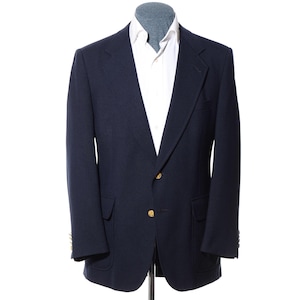 May include: A navy blue blazer featuring a notched lapel, two gold-toned metal buttons at the front closure, and two flap pockets at the waist. The blazer is layered over a crisp white collared shirt with a visible button-down placket.