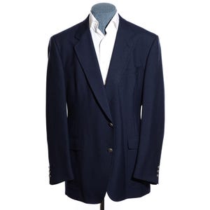 May include: A dark navy blue blazer featuring a notched lapel, two front buttons, and two flap pockets, worn over a crisp white collared shirt. The blazer has long sleeves with small buttons at the cuffs and a structured, tailored silhouette.