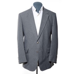 Könnte beinhalten: Ein grauer Blazer mit einer Zwei-Knopf-Vorderseite, fallendem Revers und zwei Pattentaschen an der Taille. Der Blazer wird über einem weißen Hemd mit Kragen getragen. Die Ärmel haben vier kleine Knöpfe an den Manschetten und der Stoff ist strukturiert.