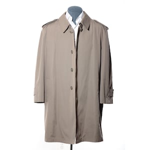 Può includere: Un trench coat beige con chiusura a bottoni, spalline e colletto a revers. Il cappotto è esposto su un manichino con una camicia bianca con colletto sotto. Il cappotto ha un design classico e su misura.