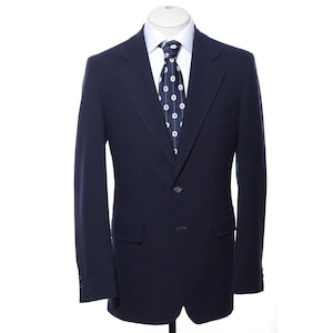 Può includere: Un blazer blu navy scuro con revers a dente, due bottoni frontali e due tasche con patta. Il blazer è indossato sopra una camicia bianca con colletto e una cravatta blu scuro caratterizzata da un motivo ripetuto di linee verticali bianche e cerchi rossi.