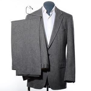 May include: A gray suit, including a blazer and trousers, displayed on a mannequin. The blazer has two buttons and a notched lapel. A white dress shirt is visible beneath the jacket. The suit is made of a textured fabric.