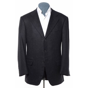Pode incluir: Um blazer cinza escuro com um padrão texturizado sutil. O blazer tem fecho de três botões, lapelas entalhadas e dois bolsos frontais. Uma camisa branca é visível por baixo. O casaco é exibido em um manequim.
